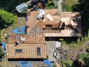 Aerial view of a house under construction with exposed framing, plywood, and scattered building materials on the roof and yard.