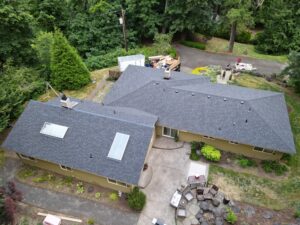 Aerial view of a suburban property with gray-roofed houses, a driveway, and a trailer loaded with lumber and construction debris beside a wooded yard.