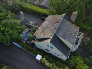 Aerial view of a suburban street with houses, a mossy roof, a black dumpster, and a tarp-covered pile in the yard.