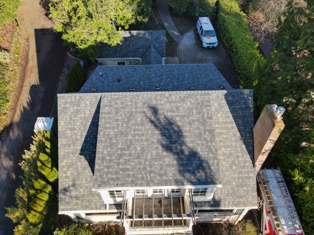 Aerial view of a gray shingled roofed house with a white balcony, curved driveway, and a white van parked among trees.