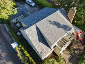 Aerial view of gray-shingle rooftops with a brick chimney and surrounding green trees, plus a white van in the driveway.