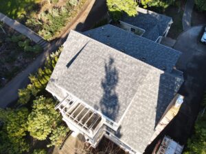 Aerial view of a large house with gray shingled roof, driveway, and surrounding trees and shrubs.