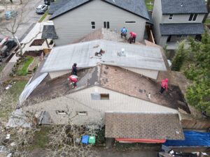 Aerial view of a residential roof repair with workers in safety gear on a brown shingle roof and scattered tools nearby.