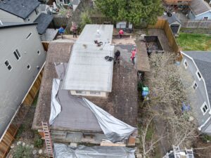 Aerial view of a backyard roof repair with workers on a flat roof, ladders, tarps, and scattered tools nearby in a suburban setting.
