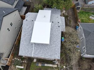 Aerial view of gray-shingled houses with a large white roof section in the center and a fenced backyard with trees.