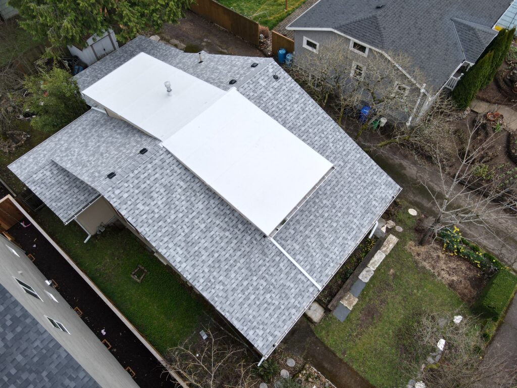 Aerial view of grey-shingled rooftops in a residential area, with a large white flat section on one roof and leafless trees nearby.