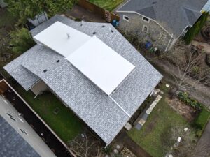 Aerial view of grey-shingled rooftops in a residential area, with a large white flat section on one roof and leafless trees nearby.