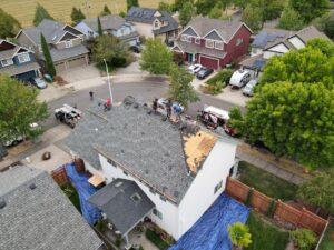 Aerial view of a two-story house roof under construction with workers on the shingles and blue tarps along the edges of the yard nearby.