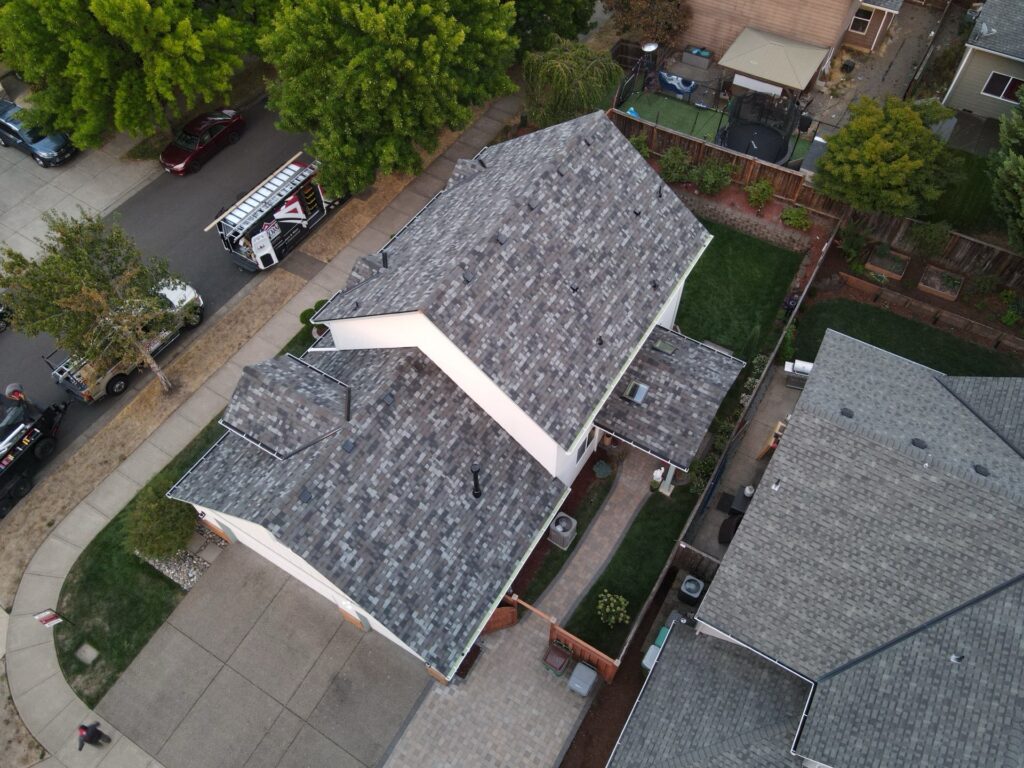 Aerial view of a suburban street with gray shingle-roofed houses, parked cars, and green yards; a backyard trampoline is visible in the upper area.