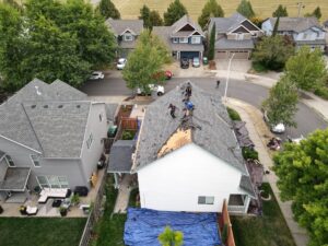 Aerial view of a suburban street with a partially reroofed house; workers stand on the roof while a blue tarp covers the ground nearby.