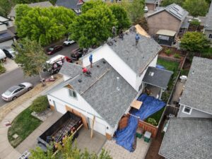 Aerial view of a house with workers on the roof installing/repairing shingles and ropes, blue tarps and ladders nearby.