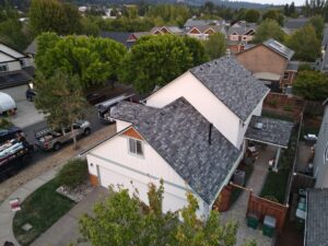 Aerial view of a suburban street with white houses, gray shingle roofs, and dense green trees lining the road.