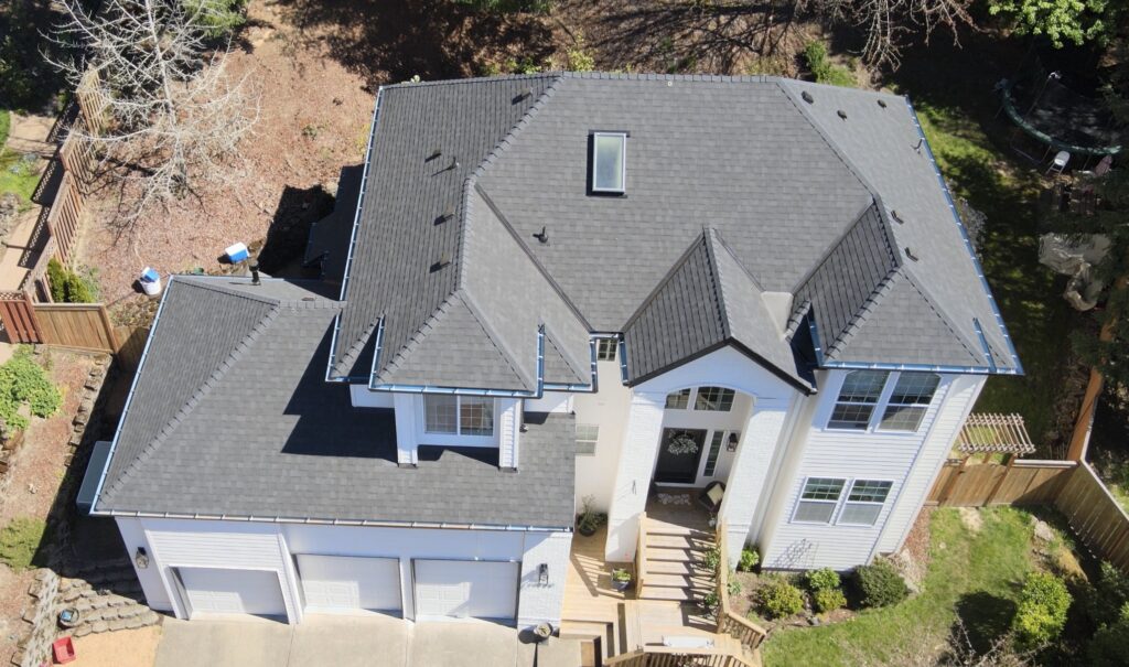 Aerial view of a two-story house with multiple gray shingled gable roofs, white exterior, wooden deck, and a trampoline in the backyard.