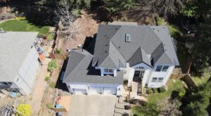 Aerial view of two houses with gray shingle roofs, backyards, and a paved driveway visible between them. A trampoline and trees surround the properties.