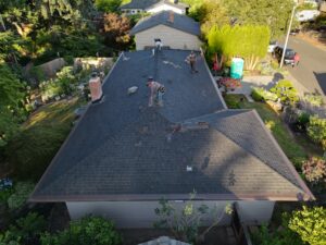 Aerial view of a house roof with multiple workers repairing shingles and tools on the roof edge, surrounded by trees and a front yard.