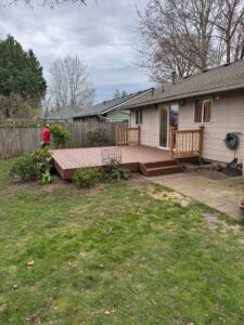Backyard with a raised wooden deck attached to a beige house, steps down to a path, and a person in a red jacket near the fence under cloudy skies.