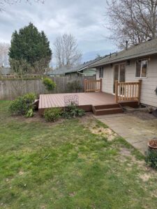 Backyard view of a raised wooden deck attached to a beige house, with stairs, railing, and a fenced yard.