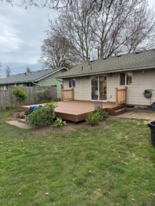 Backyard view with a wooden deck attached to a beige house, accessible by steps from a sliding glass door; small garden beds with shrubs and plants surround the deck.
