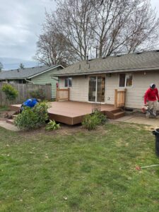 Backyard with a beige house and a new wooden deck; a worker in a red shirt operates a tool near the right side.