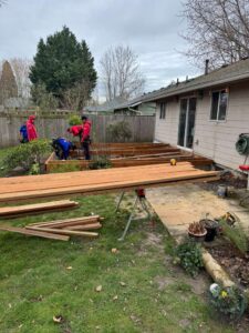 Group of workers building a wooden deck in a backyard by a beige house, with lumber and tools scattered around.