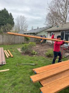 People in a backyard lift a long wooden beam while others stand nearby among lumber and tools on the grass around a house.