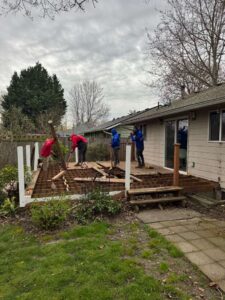 Four people install a wooden deck frame in a backyard, using tools and planks near a beige house under a cloudy sky.