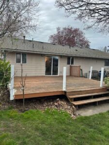 Wooden backyard deck attached to a beige single-story house, with a sliding glass door and white-post railing. Three steps lead to the yard.