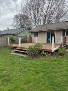 Wood deck attached to a beige house, with small steps, white posts, and surrounding shrubs in a suburban backyard.