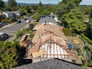 Aerial view of a tree-lined suburban street with a house under construction; workers stand on a partially built roof on a neighboring home with solar panels nearby.