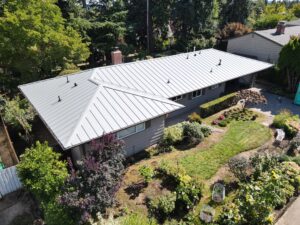 Aerial view of a single-story house with a silver metal roof, surrounded by trees and a landscaped yard.