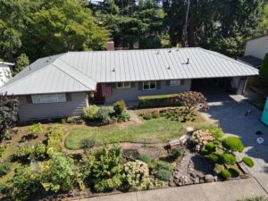 Aerial view of a single-story house with a silver metal roof, front yard garden, and driveway to the garage.