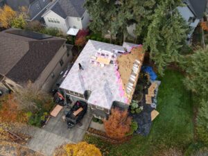 Aerial view of a house under construction with exposed roof decking and pink insulation, surrounded by trees and neighboring homes.