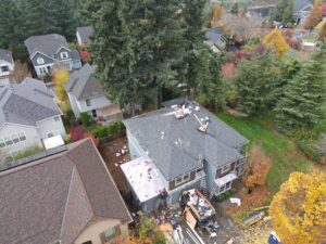 Aerial view of a suburban neighborhood with autumn trees and houses; several roofs are covered with debris and construction materials indicating ongoing renovations on the central house rooftop.