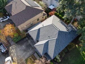 Aerial view of suburban homes with gray shingle roofs; one roof has solar panels and a white car parked in a driveway nearby.