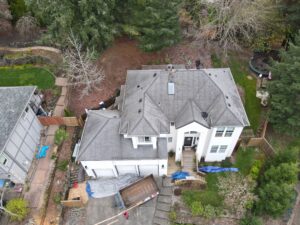 Aerial view of a two-story white house with workers on the grey shingle roof, ropes and tools nearby, and a driveway with a trailer and blue tarps in the yard.