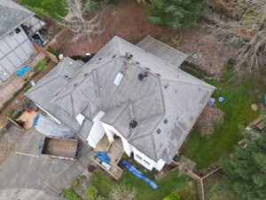 Aerial view of a house roof under repair with two workers on top, tools, ropes, and building debris strewn across the roof and yard below.
