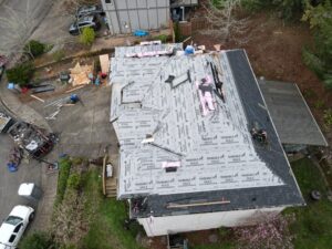 Aerial view of a house under construction with a partially covered roof and TRIBUILT MAX underlayment, scattered lumber and debris, and workers nearby in the yard.