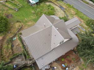 Aerial view of a large gray metal-roofed house with multiple gables, surrounded by a green yard and a road at the top edge.