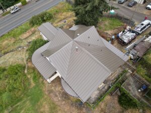 Aerial view of a large house with a multi‑gabled gray metal roof, construction equipment and a work van in the yard nearby.