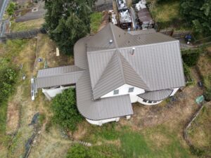 Aerial view of a house with multiple gray metal roofs, a backyard with construction materials, and surrounding trees.