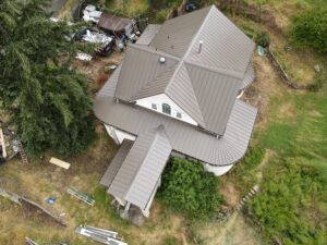Aerial view of a house with tan metal roofs, construction debris and a dumpster in the yard, two workers near material piles, and surrounding trees.
