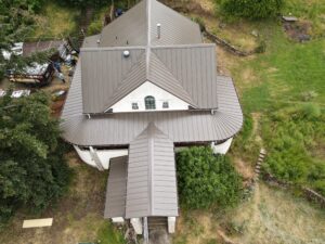 Aerial view of a multi-section metal-roof house with a cluttered work area and trailer on the left side.