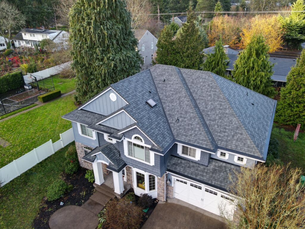 Aerial view of a blue-gray two-story house with a stone facade, circular driveway, front porch, and tall evergreen trees surrounding the property.